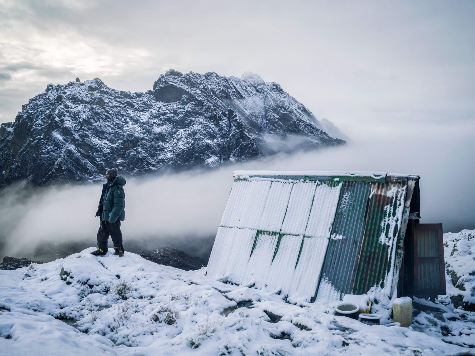 ganda Wildlife Authority Guide Muhindo Rogers stands outside Elena Hut with a view of Mt. Baker behind him. 